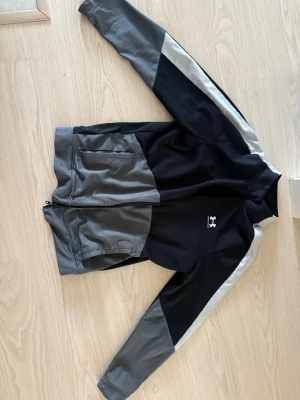 Svart och grå träningsjacka Under Armour - Sportig träningsjacka från Under Armour i svart och grått med vita detaljer. Jackan har dragkedja framtill, ståkrage och två fickor. Perfekt för träning eller uppvärmning. Materialet är troligtvis polyester för en bekväm och flexibel känsla.