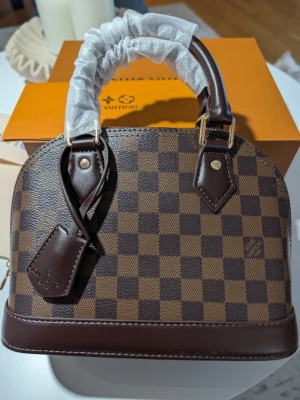 Iouis Vuitton Alma BB. - En tidlös famn av elegans Denna vackra skapelse vilar med en naturlig värdighet, klädd i ett djupt och harmoniskt schackrutigt mönster i varma toner av choklad och ebenholts. Dess mjuka, rundade former viskar om en klassisk skönhet som aldrig går ur tiden, en trofast följeslagare som bär dina hemligheter med stil.