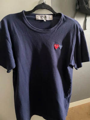 Blå Comme des Garçons Play t-shirt - Mörkblå t-shirt från Comme des Garçons Play med två broderade hjärtan på bröstet, ett rött och ett blått. Klassisk rund halsringning och korta ärmar. Tillverkad i mjuk bomull för en bekväm känsla.