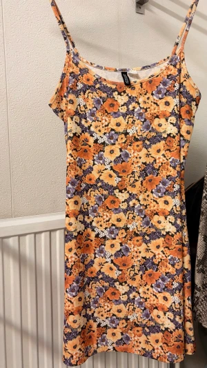 Blommig sommarklänning från HM - Söt kort klänning från HM med smala axelband och färgglatt blommönster i orange, lila och gul. Perfekt för varma dagar med sitt lätta material och avslappnade passform. Klänningen har en rak siluett och är ärmlös.