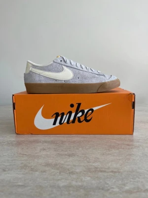 Nike Blazer Low i grå mocka - Nike Blazer Low sneakers i grå mocka med vit swoosh och vit hälpanel. Skorna har en klassisk låg siluett, vita skosnören och en brun gummisula som ger en retrovibe. Perfekta för dig som gillar stilrena och tidlösa sneakers.