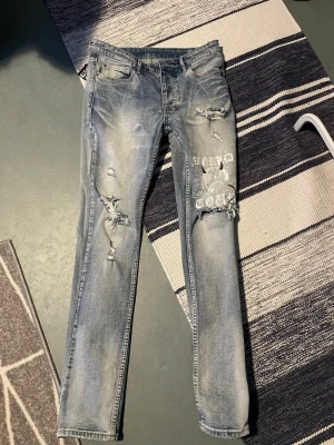 Ksubi Jeans - Säljer ett par ljusblå ksubi jeans med slitningar och tryckdetaljer på ena benet. Jeansen har en smal passform och coola trasiga partier framtill samt tryck på bakfickan. Perfekta för en trendig streetstil.