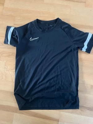 Svart Nike t-shirt med vit detalj  - Svart t-shirt från Nike med korta ärmar och vit Nike-logga på bröstet. Ärmarna har en vit rand som detalj. Perfekt för en sportig och avslappnad stil. Dri-Fit