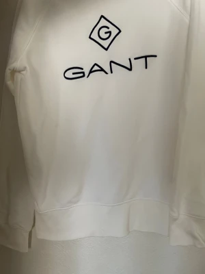 Vit sweatshirt från GANT - Snygg vit sweatshirt från GANT med broderad logga i svart på bröstet. Tröjan har rund halsringning, långa ärmar och ribbade muddar vid ärmslut och nederkant. Perfekt för en stilren och avslappnad look.