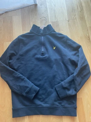 Svart Lyle & Scott tröja med dragkedja - Mörk blå långärmad tröja från Lyle & Scott med halv dragkedja och klassisk gul logga på bröstet. Tröjan har en hög krage och är tillverkad i mjukt material, perfekt för en avslappnad stil.