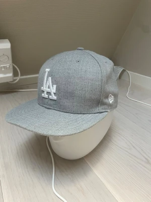 Grå LA-keps från New Era - Snygg grå keps från New Era med LA-logga broderad i vitt framtill och MLB-logga bak. Klassisk rak skärm och stilren design, perfekt för att lyfta din streetwear-look. Kepsen är gjord av 80% ull och 20% akryl. Den är använd ett fåtal gånger och är i väldigt bra skick. Storleken är 58,7 cm.