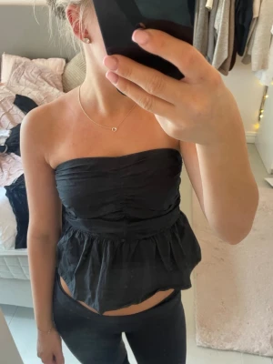 Svart axelbandslös tubtopp - Snygg svart axelbandslös tubtopp med volang nertill. Toppen har bandeau-modell och är lätt rynkad över bysten för en cool look. Perfekt att styla med jeans eller kjol för en trendig outfit. Storlek xs