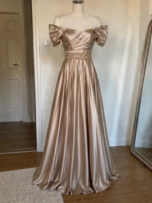 Balklänning champagnefärgad långklänning i satin oandvänd - Elegant oandvänd långklänning i champagnefärgad satin från Sword Stone. Klänningen har offshoulder-ärmar, och ett dekorerat midjeband med pärlor och paljetter. Originellt köpt för 3999kr. Fick tyvärr aldrig tillfälle att andvända den. Den har även en slits vid benet men också en underkjol. Perfekt inför en bal eller fester! 💕