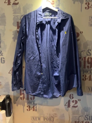 Blå skjorta från Polo Ralph Lauren - Snygg blå skjorta från Polo Ralph Lauren med klassisk krage och lång ärm. Custom fit-modell med gul broderad logga på bröstet. Perfekt för en stilren look och enkel att matcha med jeans eller chinos.
