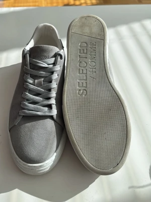Gråa sneakers från Selected Homme - Stilrena grå sneakers från Selected Homme med ovandel i mockaliknande material och vita sulor. Klassisk låg modell med vita skosnören och diskret logga på plösen. Perfekta för en clean och modern look. Helt nya använda en gång.