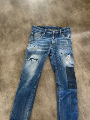 Blå jeans med patch dsquared - Säljer ett par blå jeans med slitningar och hål, samt en svart patch på höger ben. Jeansen har klassisk femficksdesign och en smal passform. Materialet är denim och de har en cool, vintage look med tydliga slitningar och detaljer.