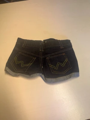 Mörkblå jeansshorts med slitningar - Snygga mörkblå jeansshorts med uppvikta benslut och slitna detaljer framtill. Shortsen har klassiska fickor, bälteshällor och kontrastsömmar i gult och orange. Bakfickorna har dekorativa sömmar i zickzack. Perfekta för en avslappnad stil.