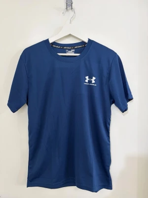 Under Armour T-shirt  - Snygg blå sport-tshirt från Under Armour med rund hals och korta ärmar. Tillverkad i ett lätt och snabbtorkande material, perfekt för träning. Logga på bröstet och i nacken ger en sportig look.