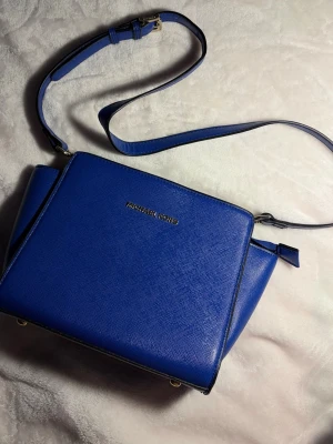 Blå axelväska från Michael Kors - Snygg blå axelväska från Michael Kors i strukturerat läder med justerbar axelrem och guldfärgad logga framtill. Väskan har en boxig form och stilrena detaljer!!