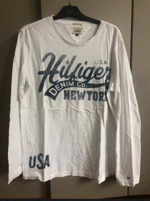 Vit långärmad Hilfiger Denim t-shirt - Vit långärmad t-shirt från Hilfiger Denim med stort blått tryck på bröstet där det står 'Hilfiger Denim Co. New York' och 'USA' nere vid nederkanten. Klassisk v-ringning och mjukt bomullsmaterial. Perfekt för dig som gillar streetstyle och amerikansk vibe.