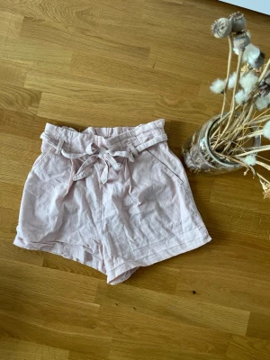 Ljusrosa shorts med knytband - Säljer ett par ljusrosa shorts med hög midja och knytband i midjan. Shortsen har avslappnad passform och är tillverkade i ett mjukt bomullstyg. Perfekta för varma sommardagar och har en söt paperbag-midja.