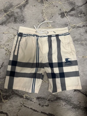 Två Burberry rutiga shorts - Snygga vita shorts från Burberry med klassiskt rutigt mönster i mörkblått . Shortsen har elastisk midja med vit snörning och Burberry-logga framtill. Perfekta för sommaren och ger en stilren look.