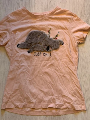 Ljusrosa t-shirt med koala-tryck - Söt ljusrosa t-shirt med ett broderat motiv av en sovande koala med renhorn och texten 'Just Chill' på bröstet. T-shirten har korta ärmar.