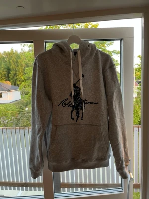 Grå hoodie från Ralph Lauren - Snygg grå hoodie, Ralph Lauren med stor svart logga och text på bröstet. Mjuk bomullsblandning och ribbade muddar vid ärmslut och nederkant. Hör av er vid frågor!