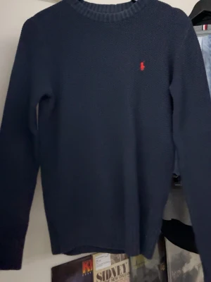 Mörkblå stickad tröja från Polo Ralph Lauren - Mörkblå stickad tröja från Polo Ralph Lauren med klassisk rund halsringning och röd broderad logotyp på bröstet. Tröjan har lång ärm och ribbade muddar vid ärmslut och nederkant. Perfekt för lager på lager och stilren look.