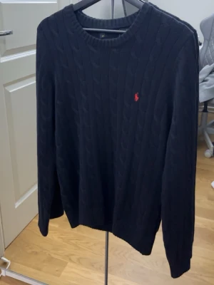 Mörkblå kabelstickad tröja Polo Ralph Lauren - Kabelstickad mörkblå tröja från Polo Ralph Lauren med rund halsringning och den klassiska röda logotypen broderad på bröstet. Långärmad modell i mjukt material, perfekt för lager på lager och en stilren look.