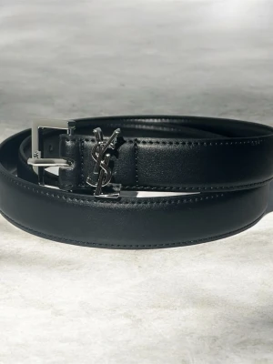 Svart skärp från Saint Laurent - Stilrent svart skärp från Saint Laurent  med silverfärgat YSL-spänne. Bältet har en klassisk design med diskreta sömmar och elegant finish. Perfekt accessoar för att lyfta din outfit.