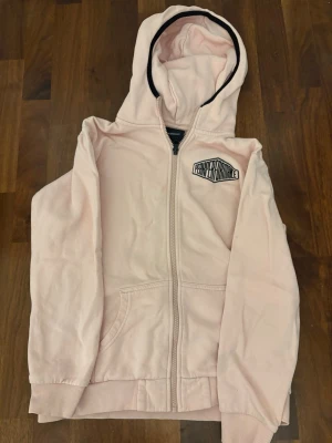 Ljusrosa zip-hoodie från Peak Performance - Snygg ljusrosa hoodie med dragkedja från Peak Performance. Hoodien har svart kant på insidan av huvan, två fickor framtill och logga på bröstet. Perfekt för en avslappnad och sportig stil. OBS! Den har nån grön fläck på armen som inte går bort.