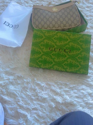 Gucci beige monogram axelremsväska - Snygg beige axelremsväska från Gucci med klassiskt GG-monogram och guldfärgade detaljer. Väskan har en elegant bågform och är tillverkad i canvas med läderdetaljer. Kommer med originalförpackning, dustbag och äkthetsintyg.