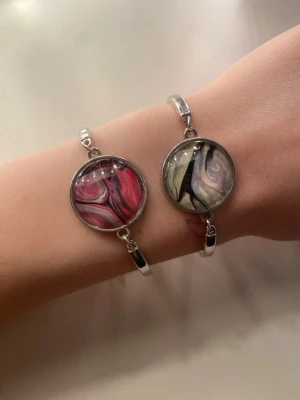 Silvriga armband med marmorerade detaljer - Två stilrena armband i silverfärgad metall med runda, marmorerade detaljer i glas. Ena armbandet har en rosa och svart swirl, det andra har en mix av svart, vitt och lila.  Helt oanvända, 10kr styck eller båda för 15kr 🫶