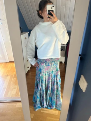Färgglad långkjol med paisleymönster - Säljer en långkjol med färgglatt paisleymönster i blått, rosa och grönt. Kjolen har smock i midjan och är tillverkad i ett lätt och luftigt material, perfekt för varmare dagar. Den har en flowy passform och ger en bohemisk vibe.
