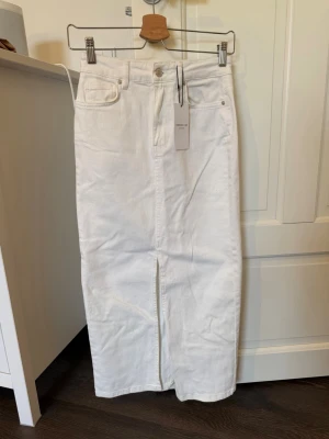 Vit långkjol i denim med slits - Snygg vit långkjol i denim från Gina Tricot med klassisk femficksdesign och slits framtill. Kjolen har hög midja och raka linjer för en clean look. Perfekt att styla med sneakers eller boots för en trendig vibe. Prislapp kvar 