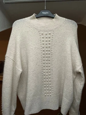 Stickad tröja med pärlor - Stickad beige tröja från KappAhl med dekorativa pärlor framtill och längs ärmarna. Tröjan har rund halsringning, ribbade muddar och en mjuk, fluffig känsla. 