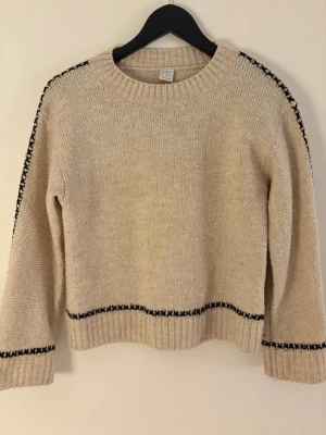 Beige stickad tröja med svarta detaljer - Beige stickad tröja med rund halsringning och svarta broderade detaljer längs ärmar, nederkant och ärmslut. Tröjan har en rak passform och är tillverkad i ett mjukt garn som känns skönt mot huden. Perfekt för lager-på-lager och enkel att matcha.
