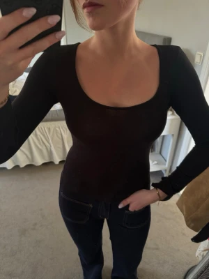 Svart långärmad topp med djup ringning - En svart långärmad topp med djup, rundad ringning. Modellen är tight och passar perfekt till jeans eller kjol. Tunt och stretchigt material som känns bekvämt mot huden. Enkel och stilren design utan mönster eller detaljer.
