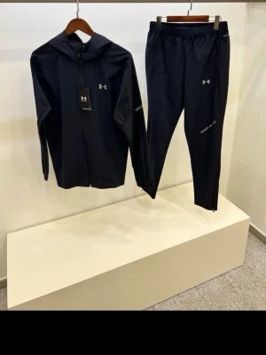 Svart träningsset från Under Armour - Snyggt svart träningsset från Under Armour med zip-hoodie och matchande byxor. Setet har diskreta loggor på bröstet och låret samt texten 'Under Armour' på ärm och ben. Perfekt för gymmet eller löprundan. Tillverkat i ett lätt och funktionellt material.