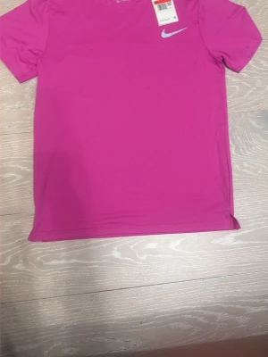 Rosa Nike t-shirt med logga - Snygg rosa t-shirt från Nike med klassisk vit logga på bröstet. T-shirten har rund halsringning och korta ärmar, perfekt för en sportig och avslappnad stil. Tillverkad i mjukt och lätt material som känns skönt mot huden.