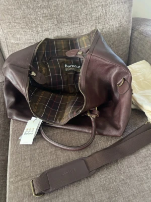 Barbour mörkbrun weekendbag i läder - Helt oanvänd!!!! Snygg och rymlig weekendbag från Barbour i mörkbrunt läder med klassiskt rutigt innerfoder. Väskan har både handtag och justerbar axelrem i matchande färg samt detaljer i mässing. Perfekt för resor när du vill ha stil och funktion. 