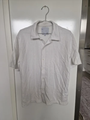 Vit kortärmad linneskjorta - Säljer en stilren vit skjorta i linne med korta ärmar och klassisk krage. Skjortan har knappar framtill och en avslappnad passform, perfekt för varma dagar. Enkel och tidlös design som passar till mycket.
