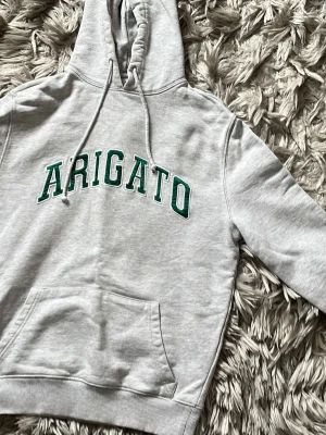 Grå Arigato hoodie med grön text - Snygg grå hoodie från Arigato med grön text på bröstet. Klassisk huva med dragsko och stor magficka framtill. Tillverkad i mjukt sweatshirtmaterial och har ribbade muddar vid ärmslut och nederkant. Perfekt för en avslappnad streetwear-look.
