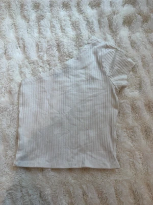 Vit ribbad topp från Gina Tricot - Snygg vit ribbad topp från Gina Tricot med kort ärm och rund halsringning. Toppen är i ett stretchigt material och har en slim passform som sitter snyggt på kroppen. Perfekt att matcha med jeans eller kjol.