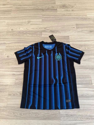 Inter Milan matchtröja 25/26 - Snygg Inter Milan matchtröja från Nike i svart med blå vertikala ränder och klubbmärke på bröstet. Tröjan har rund hals, korta ärmar och är tillverkad i ett lätt, ventilerande funktionsmaterial. Perfekt för fotboll eller som supporterplagg.