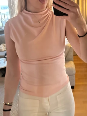 Ljusrosa ärmlös topp från Zara - Supersnygg ljusrosa topp från Zara med draperad halsringning och ärmlös design. Sitter figurnära och är gjord i ett mjukt, stretchigt material som känns skönt mot huden. Perfekt att styla med jeans eller kjol för en trendig look. Insydd lite i midjan