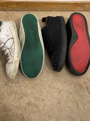 Två par sneakers, vit och svart - Säljer två par sneakers: ett par vita med gröna detaljer och ett par svarta med nitar framtill och röd sula. Båda har klassisk låg profil och snörning. Perfekta för dig som gillar unika detaljer och streetstyle.