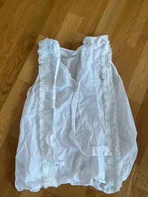 Vit ärmlös blus med spetsdetaljer - Söt vit blus med spets och volang längs framsidan. Blusen är ärmlös och har en avslappnad passform med knytband vid halsen från Zara, bra skick!