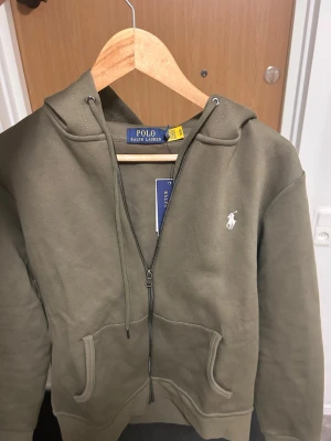 Olivgrön hoodie från Polo Ralph Lauren - Snygg olivgrön hoodie från Polo Ralph Lauren med dragkedja, huva och fickor framtill. Klassisk vit logga broderad på bröstet. Tillverkad i mjukt bomullsmaterial som känns riktigt skönt mot huden. Perfekt för en avslappnad och stilren look.