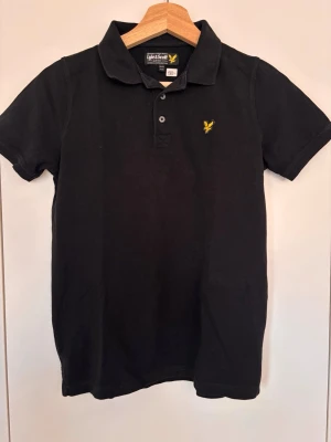 Svart pikétröja från Lyle & Scott, 12-13 år - Klassisk svart pikétröja från Lyle & Scott med korta ärmar och krage. Tröjan har två knappar framtill och den ikoniska gula loggan broderad på bröstet. Perfekt för en stilren och avslappnad look.