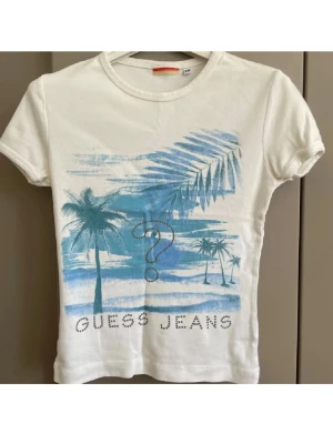 Vit t-shirt Guess Jeans med palmmotiv - Vit t-shirt från Guess Jeans med blått tryck av palmer och strand, samt ett stort frågetecken och texten 'GUESS JEANS' framtill. T-shirten har korta ärmar och rund hals. Perfekt för en avslappnad stil.