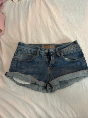 Blå jeansshorts från B.B. Jeans - Snygga blå jeansshorts från B.B. Jeans med upprullade benslut och klassisk femficksdesign. Shortsen har knappar och dragkedja framtill samt dekorativa bakfickor med lock och knapp. Perfekta för varma dagar och enkel att matcha.