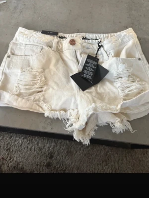 Vita slitna jeansshorts från Bershka - Säljer ett par vita jeansshorts från Bershka med slitna detaljer och fransig kant. Shortsen har klassisk femficksdesign, knapp och dragkedja framtill. Perfekta för sommaren och ger en cool, avslappnad vibe.