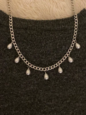 Silverfärgat halsband med pärldroppar - Stilrent halsband i silverfärgad rostfritt med sju hängande droppformade pärlor. Kedjan är grov och ger ett trendigt intryck, perfekt för att lyfta din outfit med en cool detalj. Passar dig som gillar att sticka ut med accessoarer.
